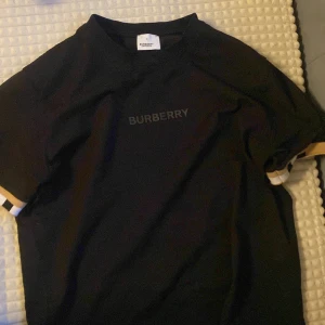 Svart t-shirt från Burberry - Säljer en stilren svart t-shirt från Burberry med logotypen tryckt på bröstet. T-shirten har korta ärmar med en vit och beige randdetalj vid ärmsluten. Perfekt för en casual look.