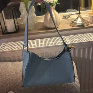 Blå axelväska med krokodilmönster - Snygg blå axelväska med krokodilmönster och dragkedja. Perfekt för att ge din outfit en trendig touch. Väskan har en stilren design och är lätt att bära med sig. Använd endast fåtal gånger. Tror den är från SHEIN men minns ej vad den kosta, runt 50-60 skulle jag tro.