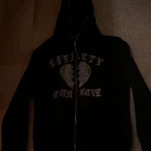 Svart hoodie med dragkedja - Säljer en svart hoodie med dragkedja och texten 'Loyalty Over Love' i ett coolt typsnitt. Designen har ett brustet hjärta i mitten. Perfekt för en avslappnad stil. Saknas några diamanter 