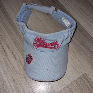 Ljusblå solskärm med rosa broderi - Snygg ljusblå solskärm med rosa broderad text 'Princess' och en liten krona. Tillverkad i bomull med justerbar rem för perfekt passform. Perfekt för soliga dagar! 54 cm, passar barn I 5-8 år.