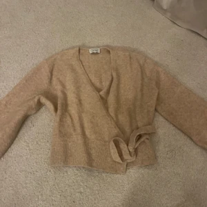 Beige kaschmir kofta från & Other Stories - Säljer en mysig och jättepopulär beige omlottkofta från & Other Stories i 100% kaschmir. Koftan har långa ärmar och ett knytband i midjan för en justerbar passform. 🫧💕