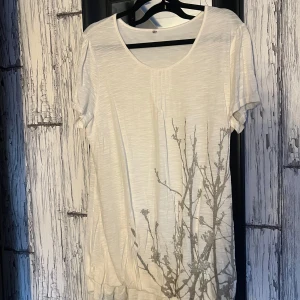 Vit kortärmad topp med tryck - Säljer en vit kortärmad topp med ett subtilt tryck av grenar på framsidan. Toppen har en rund halsringning och är tillverkad i ett lätt och bekvämt material. Perfekt för en avslappnad stil.