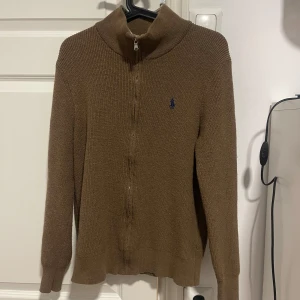 Brun stickad tröja från Ralph Lauren - Säljer en snygg brun stickad tröja från Ralph Lauren med dragkedja och långärmad design. Perfekt för kyligare dagar med sin stilrena look och bekväma passform. Den har en liten broderad logga på bröstet.