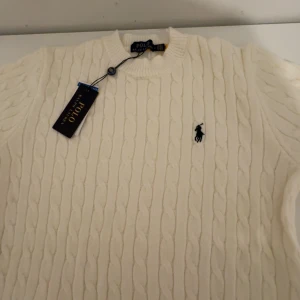 Vit stickad tröja från Polo Ralph Lauren - Säljer en stilren vit stickad tröja från Polo Ralph Lauren med det klassiska logotypbroderiet på bröstet. Tröjan har en rund halsringning och långa ärmar, perfekt för kyligare dagar.