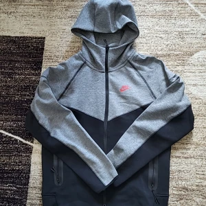 Nike tech fleece svart och grå - Säljer en snygg grå och svart nike tech fleece från Nike med dragkedja och huva. Perfekt för en sportig look. Tröjan har långa ärmar och en liten Nike-logga i rött på bröstet. Passar perfekt för träning eller en avslappnad dag.