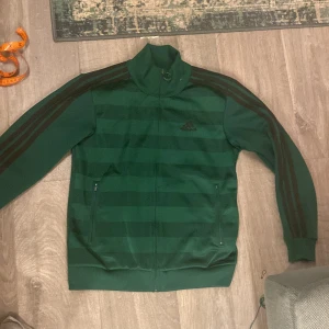 Grön träningsjacka från Adidas - Säljer en grön träningsjacka från Adidas med svarta ränder längs ärmarna och dragkedja framtill. Jackan har en hög krage och ribbade muddar vid ärmslut och nederkant. lite nopprig och använt skick men inga defekter som påverkar användning