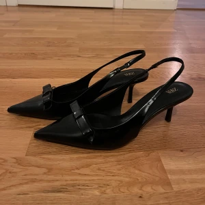 Svarta pumps från Zara - Eleganta svarta pumps från Zara med spetsig tå och en liten klack. Skorna har en stilren design med en dekorativ rosett framtill. Perfekta för en chic look.