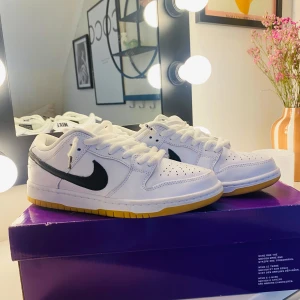Nike SB Dunk Low Pro - Säljer ett par Nike SB Dunk Low Pro i vitt med svart swoosh och gul sula. Skorna har en klassisk design med snörning och är perfekta för både stil och komfort. Säljes pga fel storlek!, pris kan diskuteras 
