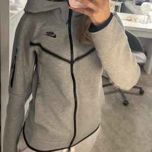 Grå huvtröja från Nike tech  - Säljer en grå huvtröja från Nike tech med dragkedja och svarta detaljer. Tröjan har en ficka med dragkedja på ärmen och är perfekt för en sportig look. Den är tillverkad i ett mjukt material och har en normal passform.