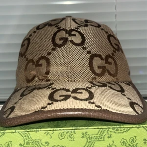 Beige keps från Gucci - Snygg beige keps från Gucci med det ikoniska GG-mönstret i brunt. Kepsen har en klassisk design med böjd skärm och justerbar passform. Perfekt för att ge en lyxig touch till din outfit.