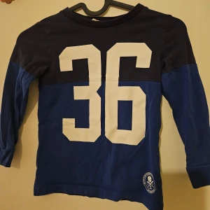 Blå och svart långärmad tröja från H&M - Säljer en cool blå och svart långärmad tröja från H&M med stort vitt nummer 36 på framsidan. Perfekt för en sportig look. Tröjan har en rund hals och är tillverkad i mjukt material för bekväm passform.