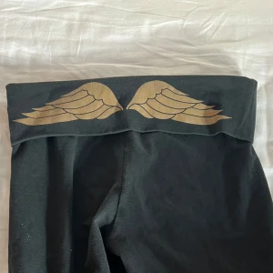 Svarta leggings med vingdetalj från Victoria's Secret - Snygga svarta från Victoria's Secret med guldiga vingdetaljer. Utsvängda bootcut. Dröm yogapantsen som inte går att köpa längre. Säljer då de är för korta för mig som är 170cm. De är stretchiga och bekväma💓