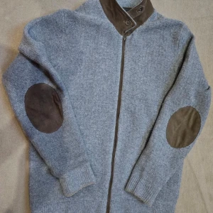 Grå ull- och kashmirtröja från Massimo Dutti - Säljer en stilren grå cardigan från Massimo Dutti i en lyxig blandning av ull och kashmir. Tröjan har en dragkedja framtill och snygga bruna armbågslappar. Tunn så passar till våren! Storlek L men närmare M i storlek. Längd mätt från hals 70 cm, bredd under armhålorna 56 cm, ärm mätt från armhålan 53 cm