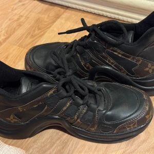 Svarta sneakers med monogramdetaljer - Snygga svarta sneakers med bruna monogramdetaljer och snörning. Skorna har en robust sula. Perfekta för en stilren look. Inte äkta 