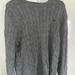 Grå stickad tröja från Ralph Lauren - Säljer en stilren grå stickad tröja från Ralph Lauren med kabelstickat mönster. Tröjan har långa ärmar och en liten röd logga på bröstet. Perfekt för kyliga dagar!