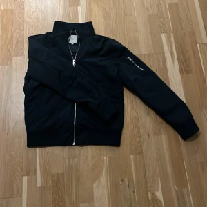 Marinblå jacka från Jack & Jones - Snygg marinblå jacka från Jack & Jones med dragkedja framtill och en ficka med dragkedja på ärmen. Perfekt för en stilren look. Jackan har en hög krage och ribbade muddar.