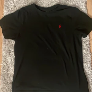 Svart t-shirt från Ralph Lauren - Säljer en klassisk svart t-shirt från Ralph Lauren med det ikoniska röda logotypen på bröstet. Perfekt för en stilren look. T-shirten är kortärmad och gjord i mjuk bomull. Passar perfekt till både jeans och shorts.