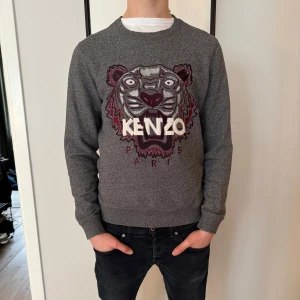 Grå tröja från Kenzo med tigerbroderi - Säljer en grå tröja från Kenzo med ett coolt tigerbroderi och texten 'KENZO PARIS' på framsidan. Tröjan har långa ärmar och en rund halsringning. Perfekt för dig som vill ha en stilren men ändå iögonfallande look. Modellen är 180 cm