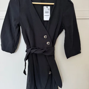 Svart klänning från Mango - Snygg svart klänning från Mango med trekvartsärmar och knappar framtill. Klänningen har en omlott-design och ett knytband i midjan för en smickrande passform.