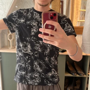 Svart t-shirt med blommigt mönster från Divided - Säljer en svart t-shirt från Divided med ett snyggt blommigt mönster i vitt. T-shirten har en bröstficka och korta ärmar, perfekt för en avslappnad stil. Passar bra till både jeans och shorts!