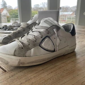 Vita sneakers från Philippe Model - Snygga vita sneakers från Philippe Model med gråa mockadetaljer och svart häl. Skorna har en klassisk design med snörning och en diskret logga på hälen. Perfekta för en stilren look.                   Den har några defekter dem är lite repiga och har gått upp lite i hälen men annars är dem felfria 