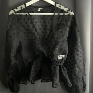 Svart prickig blus - Säljer en elegant svart blus med prickigt mönster och omlott-design. Blusen har långa ärmar och en volangdetalj i nederkant. Perfekt för en stilren look.