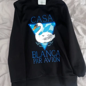 Svart sweatshirt från Casablanca - Säljer en svart sweatshirt från Casablanca med ett stort tryck av en svan och texten 'Casa Blanca Par Avion' i blått. Tröjan har en klassisk passform och är perfekt för en avslappnad stil.