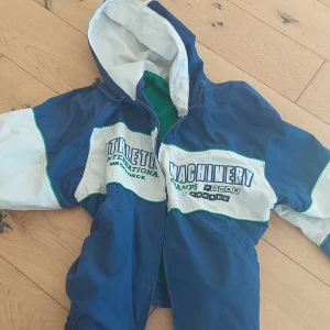 Blå och vit vindjacka med huva från 80-talet - Säljer en blå och vit vindjacka med huva och dragkedja. Jackan har tryck på framsidan och ärmarna, samt elastisk nederkant. Perfekt för blåsiga dagar!