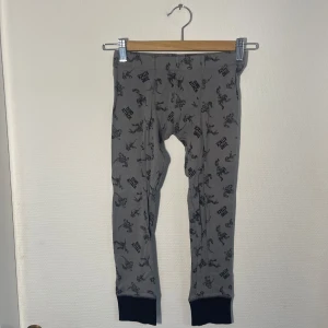 Grå leggings med dinosauriemönster från Bodyzone - Säljer ett par grå leggings från Bodyzone med ett coolt dinosauriemönster och texten 'Dino Attack'. De har svarta muddar vid bensluten och är perfekta för barn som älskar dinosaurier. Perfekta som underställ.