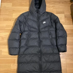 Långa Nike Jackan svart  - Säljer en svart dunjacka från Nike med huva och dragkedja. Jackan har en lång passform och är perfekt för kalla dagar. Den har två sidofickor med dragkedja och Nike-loggan på bröstet.