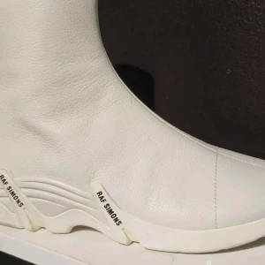 Raf Simons Cylon Sneakers, Vit, EU 42 - Helt nya kvitro finns. Stilrena vita boots från Raf Simons med en modern design. Skorna har en slät yta och en unik sula med märkets namn tryckt på sidan. Perfekta för att ge din outfit en trendig touch.
