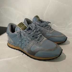 Valentino Rockrunner (passar 42,5) - Snygga Valentino Rockrunner sneakers i en stilren blå grå nyans. Skorna har en unik design med nitar på sidorna och en bekväm snörning framtill. Perfekta för att ge din outfit en lyxig touch. En nit har gått av på vänstra skon i insidan men man märken inte det. Pris kan diskuteras.