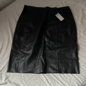 Svart pennkjol i skinnimitation - Snygg svart pennkjol i skinnimitation med hög midja och slits bak. Kjolen har en stilren och minimalistisk design som passar perfekt till många olika looks. Materialet ger en cool känsla och markerar siluetten på ett snyggt sätt.