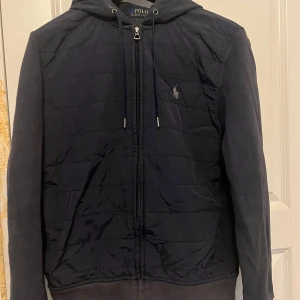 Mörkblå cardigan från Polo Ralph Lauren - Snygg mörkblå cardigan från Polo Ralph Lauren med dragkedja och broderad logga på bröstet. Jackan har quiltad framsida, huva med dragsko och långa ärmar. Perfekt för kyliga dagar.