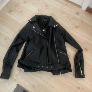 Svart skinnjacka med bälte och dragkedjor från Missguided - Säljer en svart skinnjacka med klassisk bikerstil i storlek XS. Jackan har snedställd dragkedja, silverfärgade detaljer och bälte i midjan. Flera fickor med dragkedja och långa ärmar. Perfekt för en cool och stilren look. Säljer den för att den inte har kommit till användning så mycket. 
