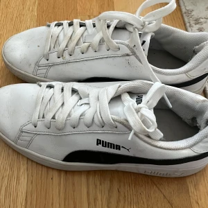 Vita sneakers från Puma - Säljer ett par klassiska vita sneakers från Puma med svarta detaljer på sidan och vit snörning. Skorna är på väldigt bra skick men är sönder vid hälen. Köpt de för 550kr och säljer de för 100kr eftersom jag inte använder de längre.
