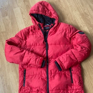 Röd pufferjacka med huva från Everest - Röd pufferjacka från Everest med svart innerfoder och huva. Jackan har dragkedja framtill och två dragkedjeförsedda fickor. Perfekt för kyligare dagar och har en sportig look.