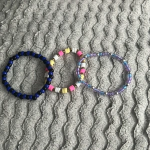 Färgglada pärlarmband - Tre handgjorda armband med pärlor i olika färger. Ett armband är mörkblått, ett har rosa, gula och vita pärlor, och det tredje har lila och blå pärlor. Perfekta accessoarer för att liva upp din stil!