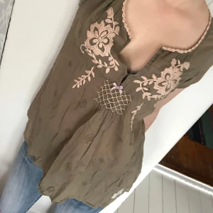 Blus med broderade detaljer från Odd Molly - Supersöt olivgrön blus från Odd Molly med ljusrosa broderade blommor och dekorativ rosett framtill. Blusen har korta ärmar, v-ringning och ett smockat parti under bysten. Perfekt för dig som gillar boho-stil!