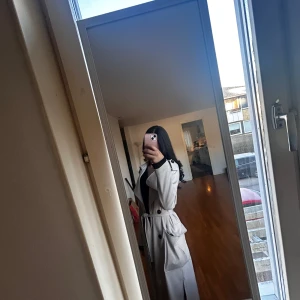 Beige trenchcoat med bälte - Snygg beige trenchcoat med klassisk krage och bälte i midjan. Jackan har långa ärmar, knappar framtill och fickor på sidorna. Perfekt för en stilren look under vår och höst.