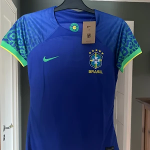 Blå Brasilien fotbollströja från Nike - Säljer en blå Brasilien fotbollströja från Nike med gula och gröna detaljer. Tröjan har korta ärmar med mönstrade partier i blått och grönt. Brasilien-logga och Nike-märke på bröstet. Perfekt för dig som vill stötta landslaget! Helt ny tags är kvar 