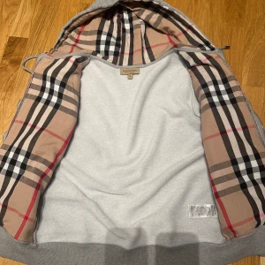 Grå hoodie från Burberry - Säljer en grå hoodie från Burberry med dragkedja och klassiskt rutigt foder i huvan. Tröjan har broderad logga på bröstet, ribbade muddar och fickor framtill. Perfekt för dig som gillar stilrena och exklusiva plagg.   !VID FRÅGOR KOM PRIVAT!
