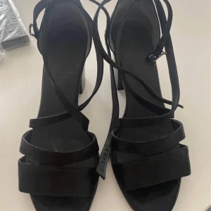 Svarta sandaler med klack och remmar - Säljer ett par svarta sandaler med bred klack och flera smala remmar som korsas över foten och runt vristen. Skorna har öppen tå och spänne vid ankeln. Perfekta för dig som vill ha en stilren och trendig look. Som ny, använd 2 gånger, klack 7, 5 cm