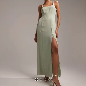 Ljusgrön långklänning med slits - Säljer en elegant ljusgrön långklänning med smala axelband och hög slits framtill. Klänningen har figurnära passform och knappar längs ryggen. Perfekt för speciella tillfällen och ger en stilren look. ALDRIG använd, med prislapp kvar!