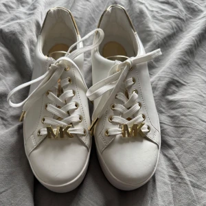 Vita sneakers  - Helt nya Snygga vita sneakers från Michael Kors (inte äkta) med guldfärgade detaljer och MK-logga framtill på snörningen. Skorna har rund tå, platt sula och guldfärgad häl. Storlek smala 36 (katt och kanin finns i hemmet) 