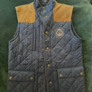 Quiltad blå väst från Lexington med mockadetaljer - Snygg quiltad blå väst från Lexington med bruna mockadetaljer på axlar och rygg. Västen har tryckknappar, två stora fickor framtill och broderad Lexington-logga på bröstet. Perfekt för lager-på-lager-stil.