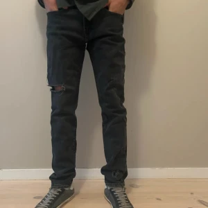 Svarta Levis 502 jeans - Säljer ett par svarta Levis 502 jeans med klassisk femficksdesign och en liten slitning på vänster knä. Jeansen har rak passform och är tillverkade i denim. 