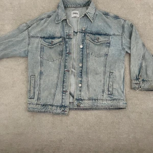 Ljusblå jeansjacka med hjärtdetalj från Zara - Säljer en ljusblå jeansjacka från Zara med en stor mörkblå hjärtformad detalj på ryggen. Jackan har klassisk krage, knappar framtill och två bröstfickor. Perfekt för dig som vill sticka ut med en unik denimlook!