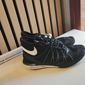 Svarta Nike sneakers med vit sula - Säljer ett par svarta Nike sneakers med vit sula och vit Nike-logga på sidan. Skorna har snörning och en sportig design med tunna linjer längs sidorna. Perfekta för träning eller vardagsbruk.