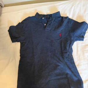 Blå pikétröja från Polo Ralph Lauren - Mörkblå pikétröja från Polo Ralph Lauren med klassisk krage och knappar framtill. Tröjan har korta ärmar och en röd broderad logga på bröstet. Perfekt för en stilren och avslappnad look. Ett litet hål men de syns inte när du använder och känns inte. Nypris runt 1.2 